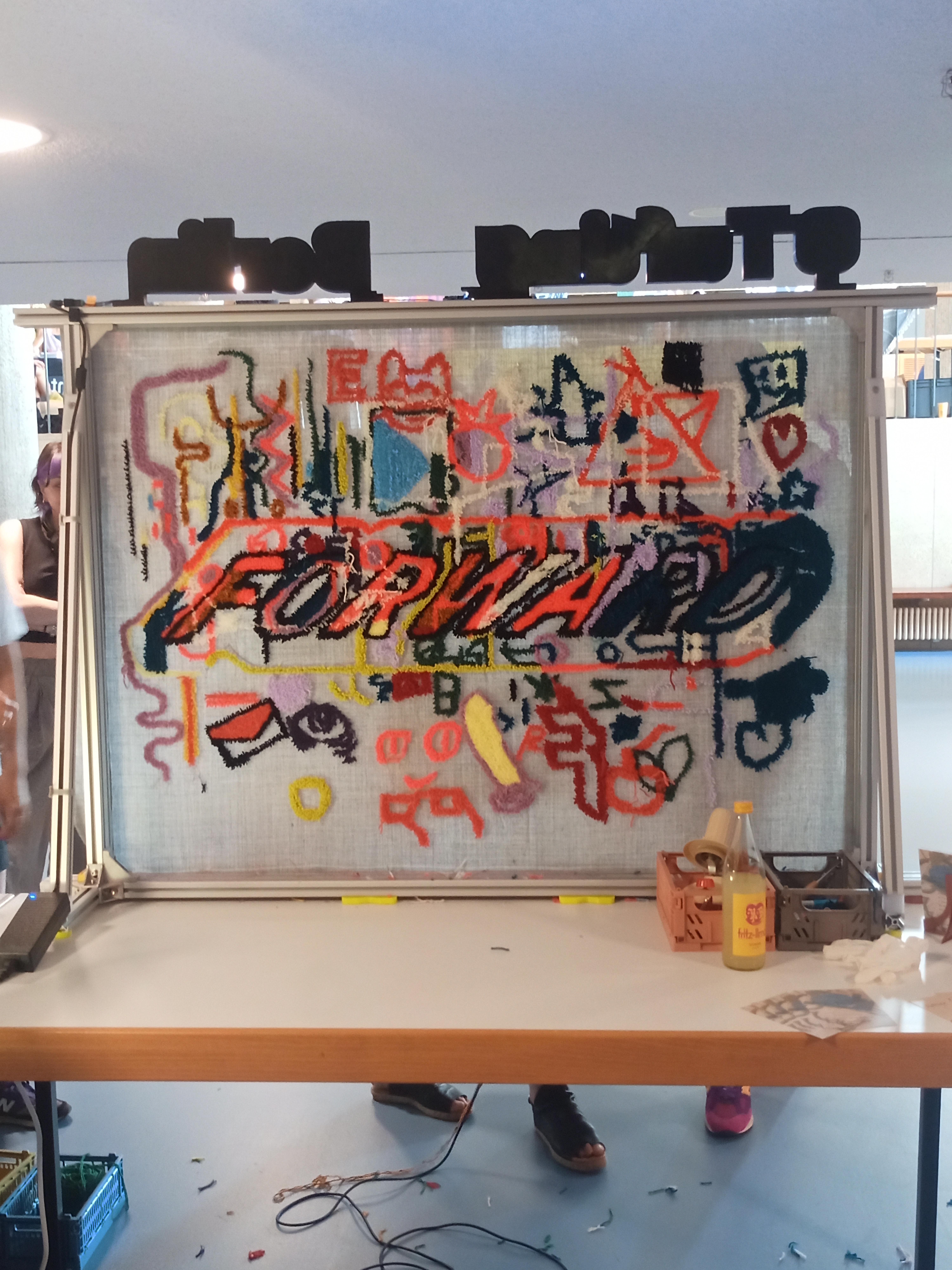 Zu sehen ist ein Foto von einem Stand beim Forward Festival im Innenbereich des HKW, wo man einen Teppich knüpfen konnte und bereits das Wort Forward geknüpft wurde